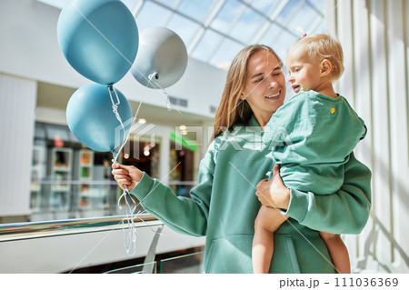 Young attractive mother with baby in her arms and colored balloons. Happy smiling mom and her little daughter in same green clothes walk around the mall and have fun. Family weekend in shopping mall. 111036369