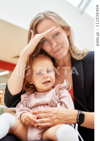 Young pretty mother with a small baby sitting on a bench in a shopping center. Mom and daughter in pink clothes hug, play, relax and have fun while shopping. Family weekend in shopping mall. 111036404