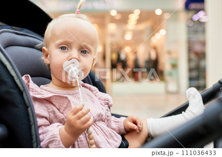 Close-up portrait of a small child in a carriage. Cute little blonde girl in pink clothes with baby pacifier. Mom with a baby in a stroller walking around the mall. Family weekend in shopping mall. 111036433