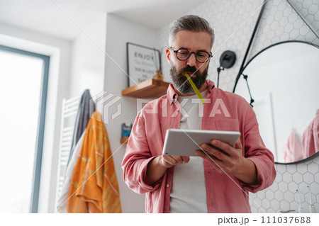 Man shopping online while brushing his teeth in bathroom. Holding tablet, scrolling and buying in eshop, toothbrush in mouth. 111037688