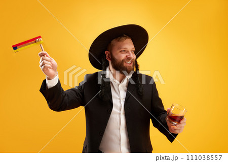 Cheerful young Jewish man in hat, with sidelocks holding noisemaker, against yellow background. Celebration 111038557
