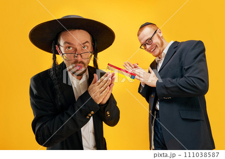 Two emotional Jewish man, friends in suits, yarmulke, with noisemaker having fun, celebrating against yellow studio background Two emotional Jewish man, friends in suits, yarmulke, with noisemaker having fun, celebrating against yellow studio background 111038587