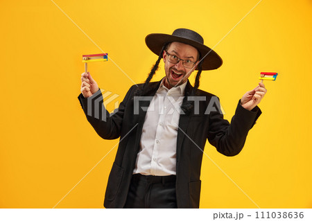 Cheerful Jewish man in hat, with sidelocks holding noisemaker, against yellow background. Celebration Cheerful Jewish man in hat, with sidelocks holding noisemaker, against yellow background. Celebration 111038636