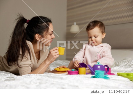 happy mother and little child daughter pretending drinking tea from plastic toy cups and spending time together in bedroom 111040217