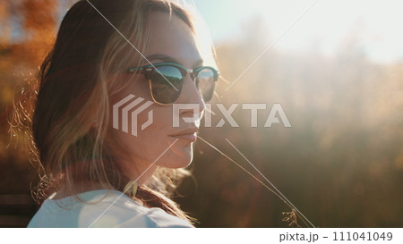 Young woman portrait in sunset soft light. Long hair caucasian girl in sunglasses holding grass blade in her mouth closeup enjoy rural forest landscape. Orange soft toning. People relax outdoor 111041049