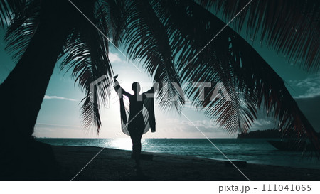 Elegant woman walk evening tropical beach under palm tree leaf. Slim joyful female silhouette raising hands, spin against sun down. Travel, relax, enjoy travel summer vacation. Blue filter back view 111041065