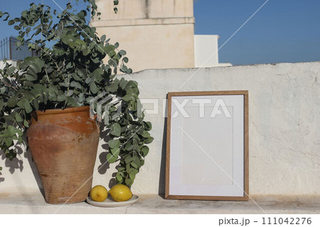 Empty vertical wooden frame picture mockup against white old textured white wall in sunlight. Fresh yellow lemons fruit, old vase with eucalyptus. Summer background, Mediterranean design. 111042276