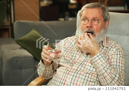 Elderly man taking pill with a glass of water, seated comfortably in a living room at home. Taking care after health 111042781
