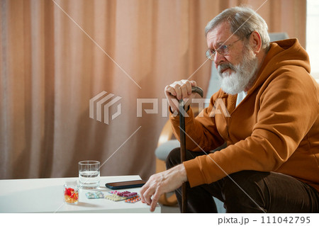 Senior man with cane sitting beside table with medications, smartphone, and a glass of water. Concentrated sad face. Meds 111042795