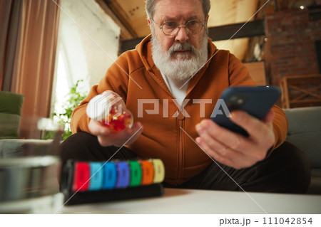 Elderly man examining pill bottle while looking at smartphone. Using pill organizer. Online med services 111042854