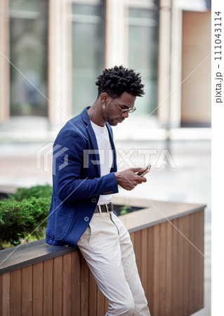 Stylish African American man in smart casual clothes, Texting on mobile phone against backdrop of city. Fashionable guy with smartphone outdoors Stylish African American man in smart casual clothes, Texting on mobile phone against backdrop of city. Fashionable guy with smartphone outdoors 111043114