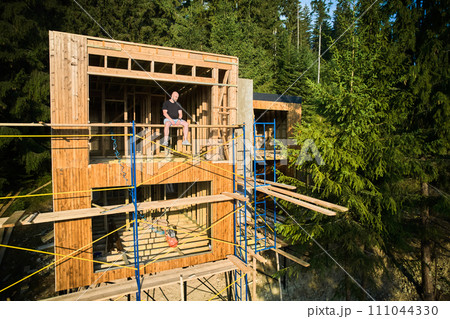 Construction manager is overseeing the building of wooden frame two-story house near the forest, sitting on scaffold. The concept behind it is modern and environmentally friendly construction. 111044330