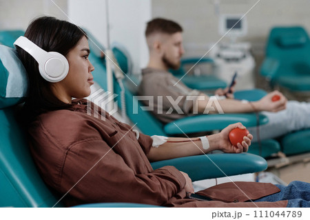 Woman listening to music sitting in armchair during transfusion, man donating blood on background Woman listening to music sitting in armchair during transfusion, man donating blood on background 111044789