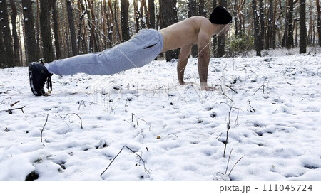 Strong man doing push ups on snow at winter forest. Sportsman exercising at nature. Young guy training outdoor. Muscular athlete showing hard and intense workout. Concept of sportive lifestyle 111045724