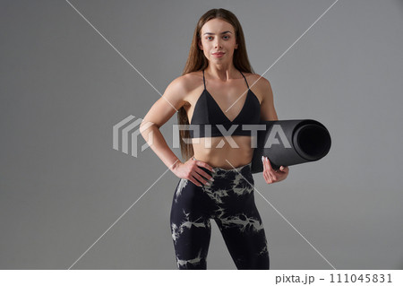 Smiling sportswoman posing with gym mat indoors. Front view of slim brunette athlete posing with hand on waist, looking at camera, during workout, isolated on grey. Sport, health care concept. Smiling sportswoman posing with gym mat indoors. Front view of slim brunette athlete posing with hand on waist, looking at camera, during workout, isolated on grey. Sport, health care concept. 111045831