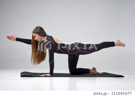 Concentrated Caucasian female practicing bird dog pose of yoga on gym mat indoors. Side view of long-hared woman in black apparel performing Parsva Balasana, isolated on grey background. Yoga concept. Concentrated Caucasian female practicing bird dog pose of yoga on gym mat indoors. Side view of long-hared woman in black apparel performing Parsva Balasana, isolated on grey background. Yoga concept. 111045877