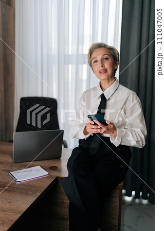 Vertical portrait of successful middle-aged business woman ceo executive manager holding smartphone, using mobile apps on cellphone, sitting on work desk in office, smiling looking at camera. 111047005