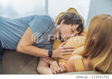Tender moment as husband lovingly embraces his pregnant wife's belly while they lie together on the sofa, sharing warmth and anticipation 111052781