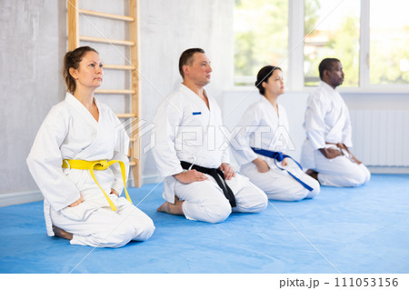 Multinational athletes dressed in white kimonos after active workout sit on floor in row and meditate 111053156