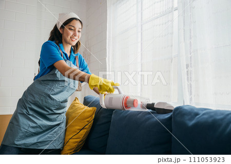 In a comfortable living room an Asian woman maid uses a vacuum machine cleaner to clean a sofa. Her dedication to hygiene and furniture care reflects modern approaches to housework. In a comfortable living room an Asian woman maid uses a vacuum machine cleaner to clean a sofa. Her dedication to hygiene and furniture care reflects modern approaches to housework. 111053923