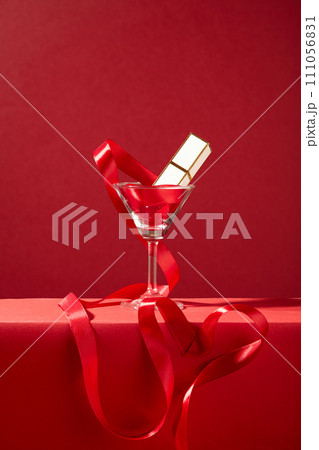 A lipstick and red silk ribbon inside a cocktail glass on a red background. Romantic gift for Valentine Day. The space is viewed from the front for advertising. 111056831