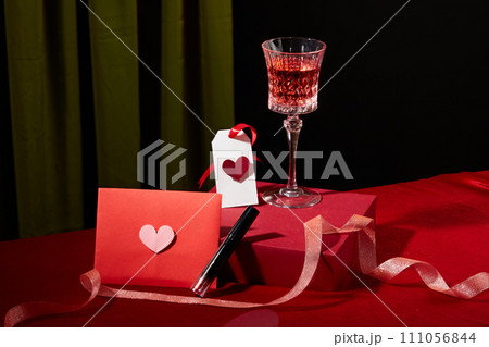 A glass of red wine is placed on a red podium, next to a lipstick, a red envelope with a pink paper heart and decorative ribbon. Romantic atmosphere. A glass of red wine is placed on a red podium, next to a lipstick, a red envelope with a pink paper heart and decorative ribbon. Romantic atmosphere. 111056844