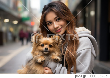 A young woman of Asian appearance holds a Spitz dog in her arms while walking on a city street 111062503