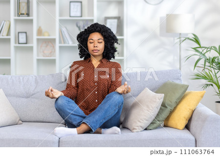 Concentrated and calm African American woman sitting on sofa at home in lotus position, doing yoga and meditating with eyes closed. Concentrated and calm African American woman sitting on sofa at home in lotus position, doing yoga and meditating with eyes closed. 111063764