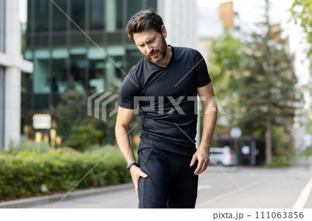 A young male runner stands outside on the street and holds his leg, pulled a muscle, gives a massage, suffers from pain. 111063856