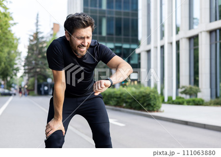 Young male athlete standing outside on elbow bent after jogging and marathon and looking disappointed and upset at smart watch on wrist, not happy with result. 111063880