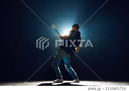 Performer with electric guitar leaning into musical moment against dark background with spotlights behind him. Performer with electric guitar leaning into musical moment against dark background with spotlights behind him. 111065718
