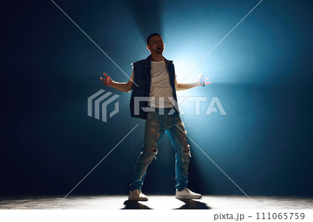 Casual dressed male singer with microphone intense performance under spotlight in darkness in dramatic smoke on stage. Casual dressed male singer with microphone intense performance under spotlight in darkness in dramatic smoke on stage. 111065759