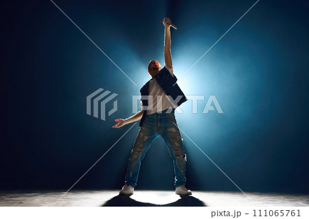 Street style singer gesturing with hand on stage against dark background. Spotlight and smoke creating silhouette effect. 111065761