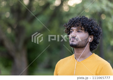 An Indian sportsman with earphones takes a break in the park. Fitness, wellbeing, and an active lifestyle captured in a tranquil outdoor setting. An Indian sportsman with earphones takes a break in the park. Fitness, wellbeing, and an active lifestyle captured in a tranquil outdoor setting. 111065777