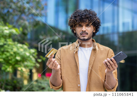 Close-up portrait of a young Indian man standing outside on the street, holding a mobile phone and a credit card, looking worriedly at the camera. 111065841