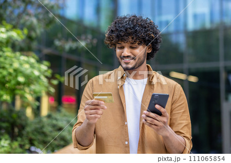 Indian smiling young man standing on city street, holding phone, looking and using credit card. 111065854