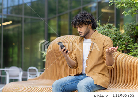 A young Indian man sits on a bench outside and looks worriedly at the phone screen, throws up his hands in frustration, received bad news by message. A young Indian man sits on a bench outside and looks worriedly at the phone screen, throws up his hands in frustration, received bad news by message. 111065855