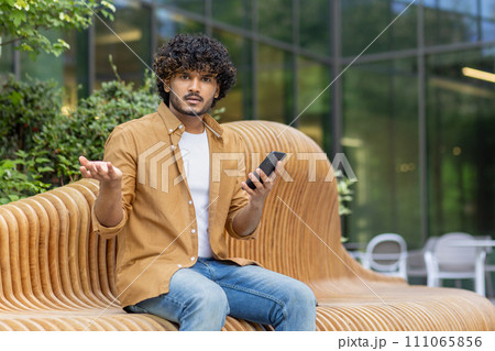 Portrait of a sad young Muslim man holding a phone in his hands, having received bad news, worriedly spreading his hands to the camera while sitting on a bench on the street. 111065856