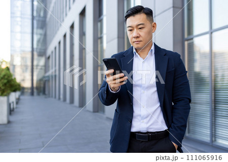 Serious young Asian man standing outside an office building in a suit, holding his hand in his pocket and using a mobile phone. 111065916
