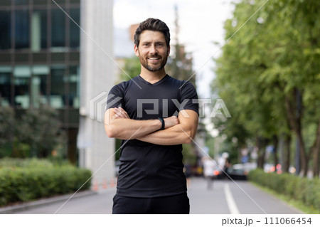 Portrait of a young sportsman standing on a city street, arms crossed on his chest, confidently and smilingly looking at the camera. Portrait of a young sportsman standing on a city street, arms crossed on his chest, confidently and smilingly looking at the camera. 111066454
