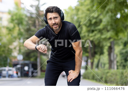 Portrait of a tired young man standing on a city street, looking at the camera and checking the result after a run on a smart watch. Portrait of a tired young man standing on a city street, looking at the camera and checking the result after a run on a smart watch. 111066476