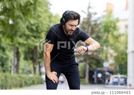 Tired young male sportsman wearing headphones standing outside in park, frowning and looking unhappily at wrist watch. 111066483