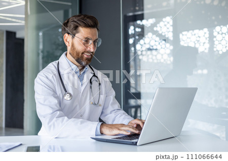 A smiling young male doctor is working in the office of the clinic using a laptop. Sitting in a white coat at the desk, typing, texting. Close-up photo. A smiling young male doctor is working in the office of the clinic using a laptop. Sitting in a white coat at the desk, typing, texting. Close-up photo. 111066644