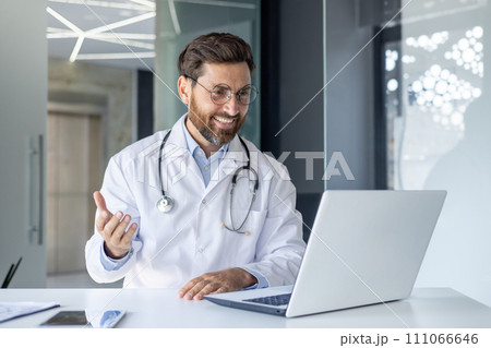 A smiling young male doctor sits in the office of the clinic at a desk, communicates and consults online through a laptop. 111066646