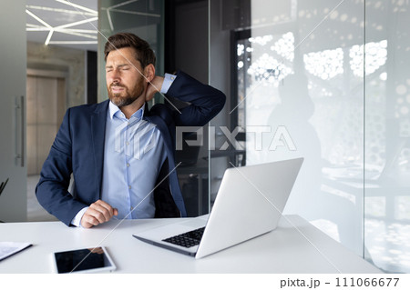 Tired young businessman working in the office using a laptop, sitting at the table and holding his neck, grimacing in pain, doing a massage. Tired young businessman working in the office using a laptop, sitting at the table and holding his neck, grimacing in pain, doing a massage. 111066677