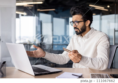Frustrated and worried young muslim man sitting in office and looking at laptop screen, holding credit card in hand and waving hands in confusion. 111067662