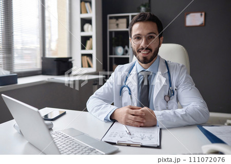 Portrait of cheerful bearded Muslim man physician wearing eyeglasses looking at camera sitting at office desk in modern clinic 111072076