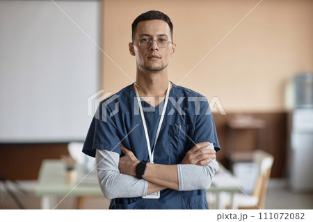 Waist up shot of confident Muslim man doctor in glasses and blue scrubs looking at camera while standing with arms crossed at chest in clinic 111072082
