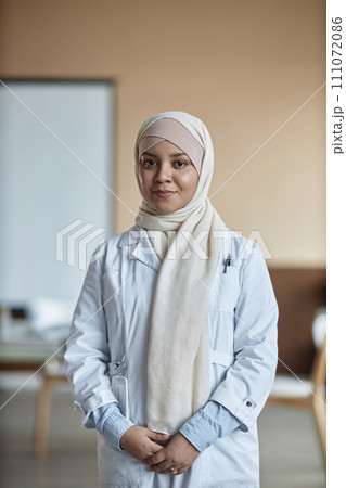 Vertical shot of smiling Islamic woman doctor in cream hijab and lab coat looking at camera standing with arms at stomach in hospital 111072086