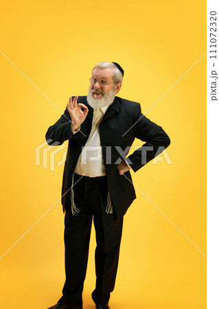 Portrait of smiling mature man in suit and black kippah making positive hand gesture against sunny yellow background. Happy Purim. 111072320